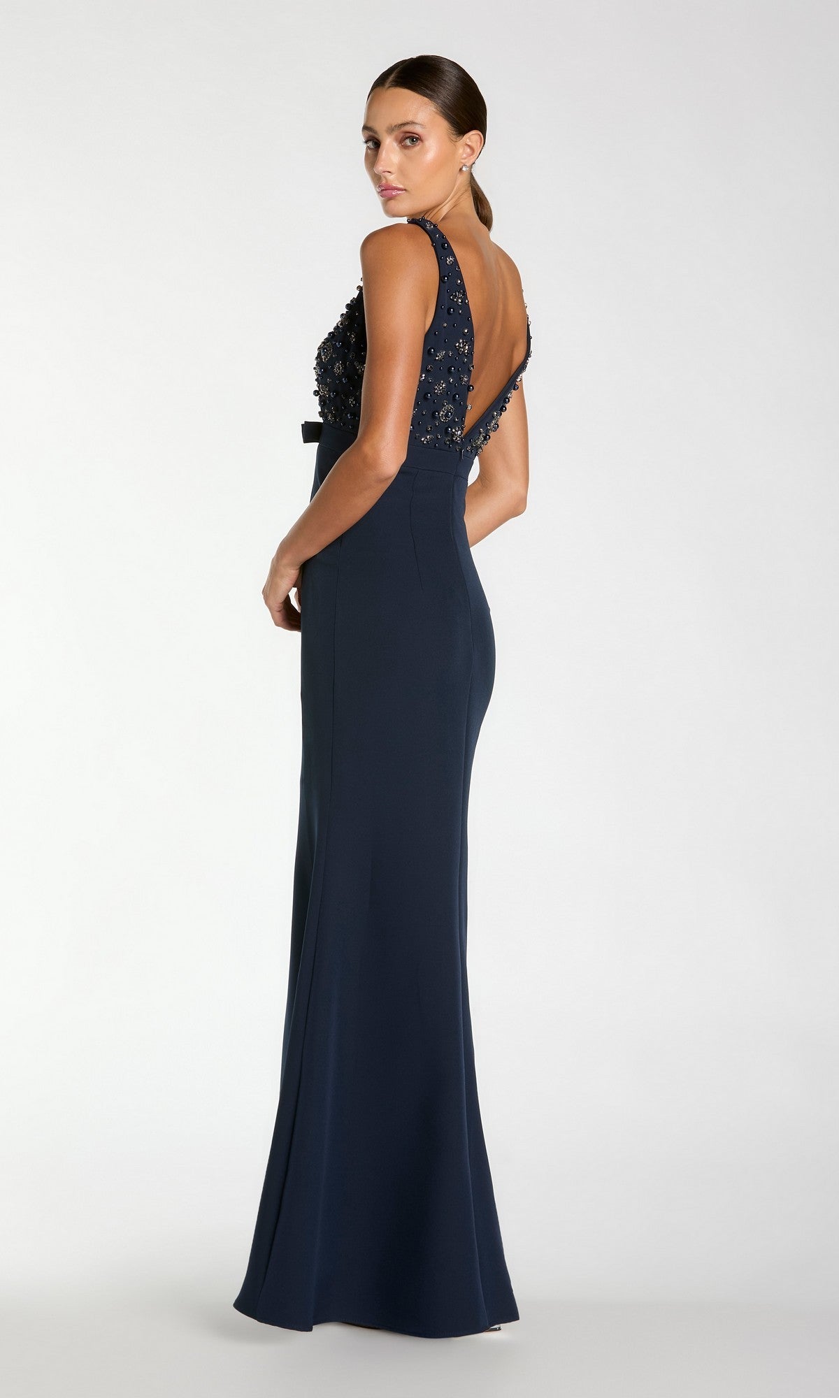 A woman wears the Beaded-Bodice Long Prom Dress by Mac Duggal 2278, featuring a floor-length navy skirt, beaded bodice, and open back as she stands facing away, looking over her shoulder.